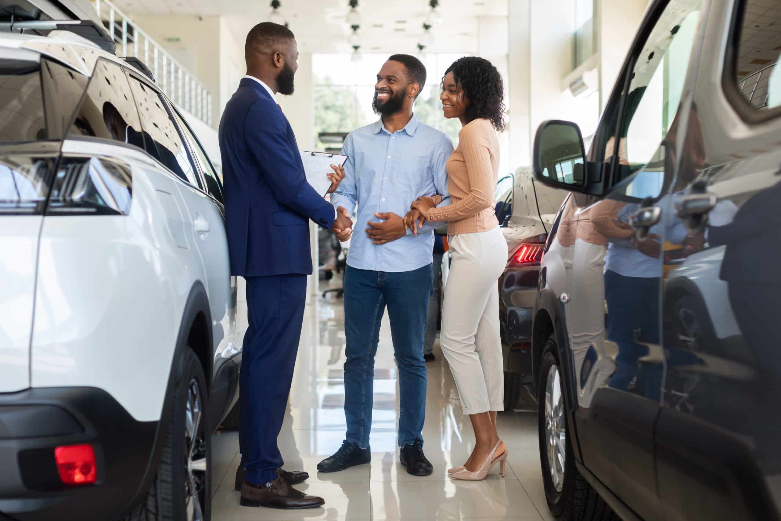 A smiling man and woman stand between vehicles in a car dealership as the man shakes hands with a car salesperson.