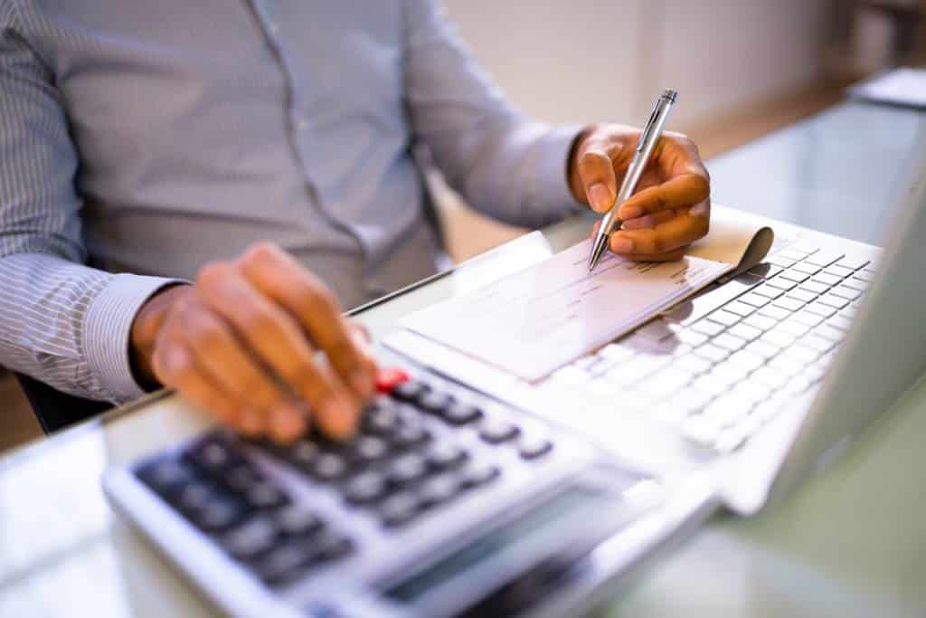 Man sitting at desk figuring payroll with pen, paycheck, laptop, and calculator