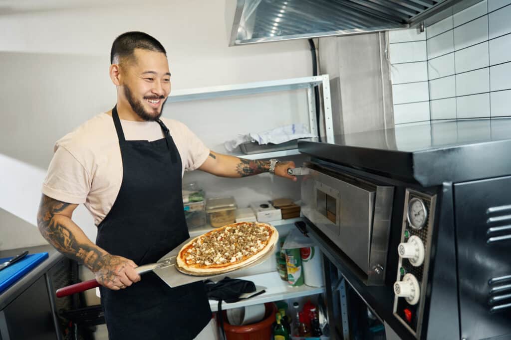 Smiling man wearing an apron prepares to slide a pizza into a restaurant oven.